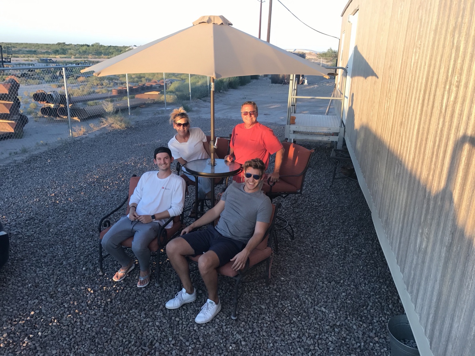 Family seated around a patio table under an umbrella at an oil field yard outside Pecos, Texas, with pipe racks and a field office trailer behind them