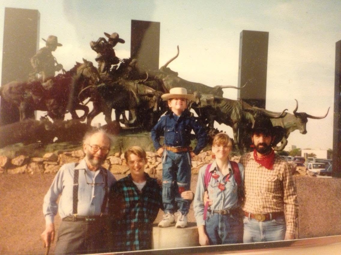 Family photo from the early 1990s in front of a bronze longhorn cattle-drive sculpture, with a young boy in a straw cowboy hat standing on the pedestal